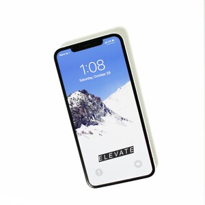 以下が含まれることがあります： 黒い画面に1:08と10月26日土曜日が表示されている白いiPhone。背景画像は雪山。画面に黒い文字で「ELEVATE」と表示されている。