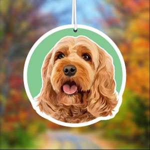 May include: A round car air freshener with a green background and a close-up image of a brown and gold dog with its tongue sticking out.