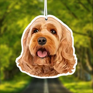 May include: A car air freshener featuring a cartoon illustration of a golden brown dog with floppy ears. The dog has a pink tongue sticking out and is looking directly at the viewer.