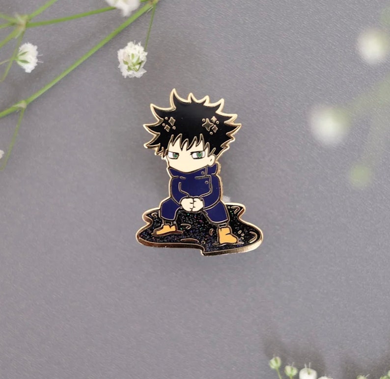 Pode incluir: Um broche de esmalte dourado com um personagem de desenho animado com cabelo preto, uma jaqueta azul e botas laranja. O personagem est&aacute; sentado em uma po&ccedil;a de glitter preto.