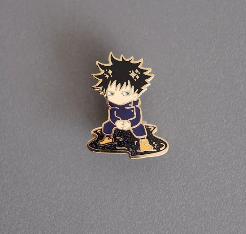 Pode incluir: Um broche de esmalte dourado com um personagem de desenho animado com cabelo preto, camisa azul e botas amarelas. O personagem est&aacute; sentado em uma po&ccedil;a preta e dourada.