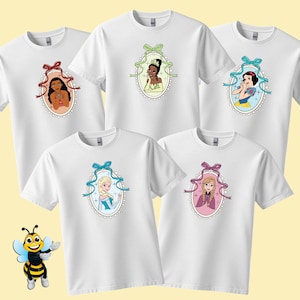 Puede incluir: Camisetas blancas con retratos de dibujos animados de princesas de Disney en marcos ovalados. Cada camiseta muestra una princesa diferente, enmarcada por una cinta y un borde de encaje. Las camisetas se muestran sobre un fondo amarillo claro.