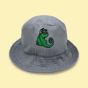 May include: A gray bucket hat featuring an embroidered green chameleon with a red tongue. The hat has a wide brim and a rounded crown. The chameleon is in a cartoon style.