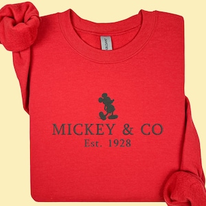 May include: A red sweatshirt with the text "MICKEY & CO Est. 1928" in black. The design includes a silhouette of Mickey Mouse. The sleeves are folded and arranged on the sides.
