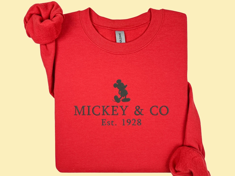 May include: A red sweatshirt with the text "MICKEY & CO Est. 1928" in black. The design includes a silhouette of Mickey Mouse. The sleeves are folded and arranged on the sides.