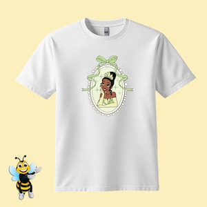 Puede incluir: Camiseta blanca con una ilustraci&oacute;n de dibujos animados de una princesa con un vestido y corona verdes, enmarcada por una cinta verde y un borde de perlas. El dise&ntilde;o est&aacute; centrado en la camiseta, ofreciendo una est&eacute;tica caprichosa y colorida.