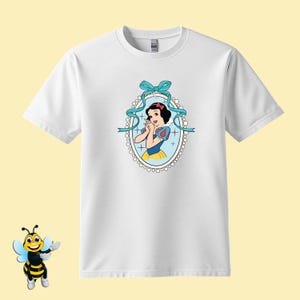 Puede incluir: Camiseta blanca con una imagen de dibujos animados de Blancanieves en un marco ovalado azul y blanco con un lazo azul. La imagen de Blancanieves tiene un bob negro, un lazo rojo y un vestido azul y amarillo.