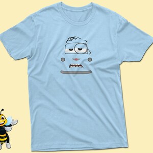 May include: Light blue t-shirt with a cartoon image of a blue car with a face. The car has a single eye, a mouth, and a cracked windshield.