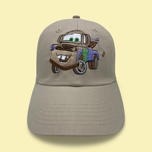 May include: A tan baseball cap featuring an embroidered image of Tow Mater from the movie Cars. The image is detailed with brown, blue, green, and white thread. The cap has a curved brim and a structured crown.