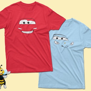 May include: Two short-sleeved t-shirts, one red and one light blue, each featuring a cartoon car face design. The red shirt has a smiling car face, while the blue shirt has a different car face design. Both shirts are on a light yellow background.