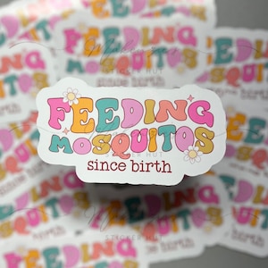 May include: A white sticker with the text "FEEDING MOSQUITOS since birth" in a retro font. The word "FEEDING" is in orange, "MOSQUITOS" is in teal and pink, and "since birth" is in brown. A daisy is on the sticker.