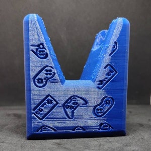 May include: A blue 3D-printed phone stand with a V-shape design. The stand is decorated with multiple game controller icons. The stand is designed to hold a phone or tablet.
