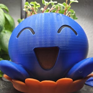 May include: A blue, smiling, spherical planter with a wide open mouth and two curved eye cutouts. The planter has orange petal-like features at the base and sprouts of green plants on top. The planter is a unique decorative item.