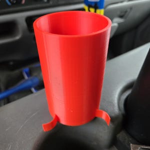 May include: A bright red, cylindrical cup with two small, curved tabs at the base. The cup is made of a solid material and appears to be 3D-printed. It is sitting on a gray surface, possibly inside a vehicle.
