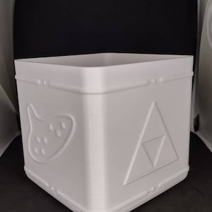 May include: A white, square-shaped container with embossed designs. One side features a stylized ocarina, while another displays a triangle design. The container has a decorative border and is likely 3D-printed.