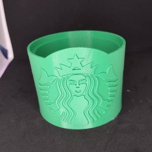 May include: A green 3D-printed cup sleeve featuring the Starbucks logo. The sleeve is cylindrical with a detailed embossed design of the Starbucks siren logo. The sleeve is designed to fit around a standard coffee cup.