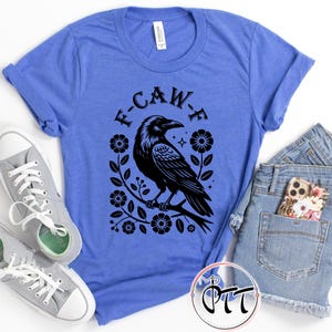 Op de afbeelding: Een blauw t-shirt met een zwarte ravenafbeelding, omringd door bloemen en de tekst "F-CAW-F". Het shirt wordt gecombineerd met grijze sneakers en een spijkerbroek. Het shirt is gemaakt van een zacht materiaal.