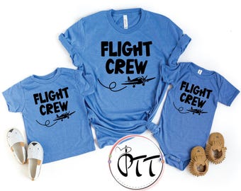 May include: Three light blue t-shirts and a onesie with the words "FLIGHT CREW" in bold black letters. Each shirt features a black airplane graphic with a curved trail. The shirts are accompanied by a pair of baby shoes and slippers.