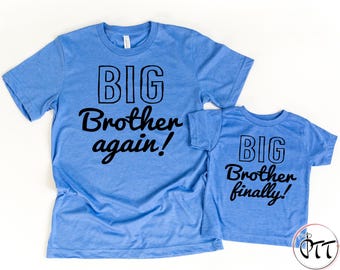 May include: Two light blue t-shirts with black text. The larger shirt reads "BIG Brother again!" and the smaller shirt says "BIG Brother finally!" Both shirts have short sleeves and a crew neck.