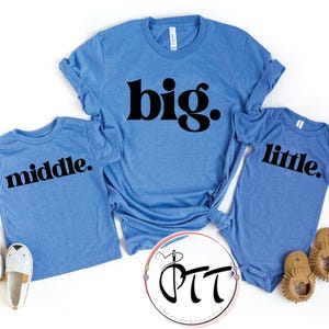 May include: Three light blue shirts with black text that reads "big.", "middle.", and "little." The shirts are accompanied by a pair of white and gray slip-on shoes and a pair of brown baby moccasins. The shirts are likely family matching apparel.