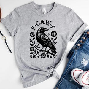 Op de afbeelding: Een lichtgrijs t-shirt met een zwarte raaf op een tak, omringd door bloemmotieven. De tekst "F-CAW-F" staat erboven. Het shirt wordt gecombineerd met een blauwe jeans en witte sneakers.