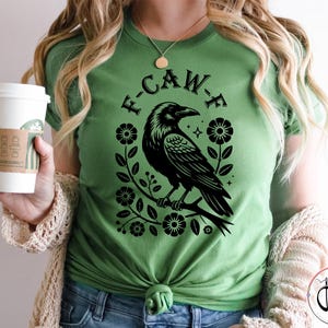 Op de afbeelding: Groen t-shirt met een zwarte raven illustratie en de tekst "F-CAW-F". De raaf zit op een tak, omringd door bloemmotieven. Het t-shirt heeft een effen groene kleur.