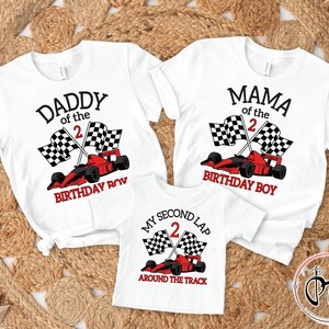 May include: Three white t-shirts featuring a racing theme. The shirts display red race cars, checkered flags, and the text DADDY OF THE 2 BIRTHDAY BOY, MAMA OF THE 2 BIRTHDAY BOY, and MY SECOND LAP 2 AROUND THE TRACK in black and red lettering.