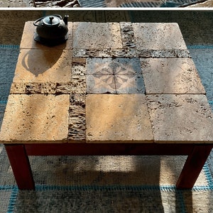 Puede incluir: Una mesa de centro cuadrada hecha de piedra natural con una superficie rústica y texturizada. La superficie de la mesa está compuesta por múltiples azulejos con un patrón geométrico. La mesa tiene un marco de madera oscura y se encuentra sobre una alfombra estampada.