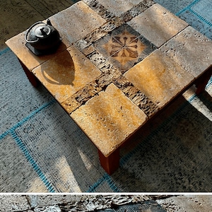 Op de afbeelding: Een vierkante salontafel gemaakt van beige en bruine tegels met een zwart-wit bloemenmotief in het midden. De tafel heeft een rustieke, landelijke stijl en staat op een blauw-wit tapijt.