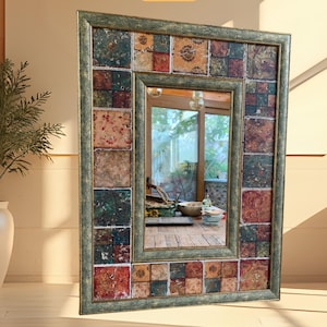 Puede incluir: Espejo rectangular con marco de mosaico de azulejos en tonos verdes, rojos y marrones. El espejo refleja una habitación con una mesa de madera y una planta en una maceta beige. El marco tiene un aspecto envejecido.