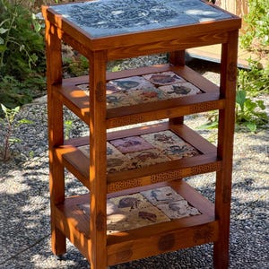 Può includere: Un tavolino in legno con tre ripiani, ciascuno con piastrelle decorative. La piastrella superiore ha un design blu e bianco, mentre i ripiani inferiori mostrano piastrelle colorate con vari motivi. Il tavolo è tinto di un marrone caldo.