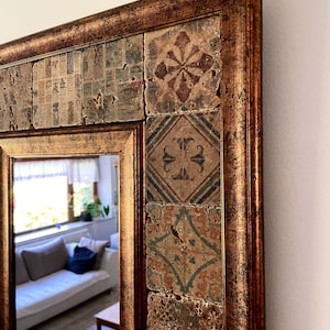 May include: A decorative mirror with a rectangular frame. The frame has a distressed gold finish. The mirror's border is adorned with square tiles featuring various patterns in shades of brown, green, and blue. The mirror reflects a room with a white couch.