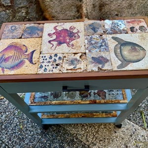 May include: A blue and brown serving cart with a tiled top featuring various sea creature illustrations. The cart has a drawer with a black handle and a lower shelf. The frame is light blue, and the top is framed in brown wood.