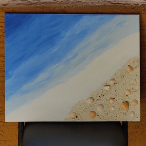 May include: A blue and white canvas painting depicting a beach scene with seashells scattered on the sand.