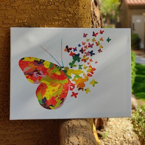 May include: A white canvas featuring a colorful butterfly design. The butterfly's body is a mix of red, yellow, and green, with smaller butterflies in various colors trailing off. The artwork is displayed outdoors.