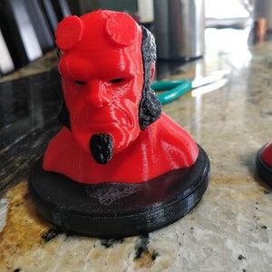 May include: A red 3D printed bust of a character with horns, a black beard, and black hair. The bust is on a black base.