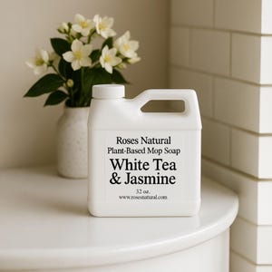 May include: A white plastic bottle of Roses Natural Plant-Based Mop Soap. The label reads "White Tea & Jasmine" and "32 oz." A vase of white flowers sits in the background. The website www.rosesnatural.com is also listed on the label.