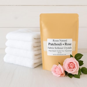 May include: A stack of four white towels next to a tan pouch of fabric softener crystals. The pouch has a label that reads "Roses Natural Patchouli + Rose Fabric Softener Crystals." Two pink roses with green leaves are in front of the pouch.