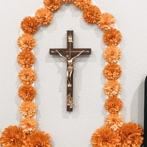 Peut inclure: Un mur blanc avec une croix en bois et une figurine dor&eacute;e de J&eacute;sus-Christ. La croix est entour&eacute;e d'une guirlande de fleurs en papier orange.