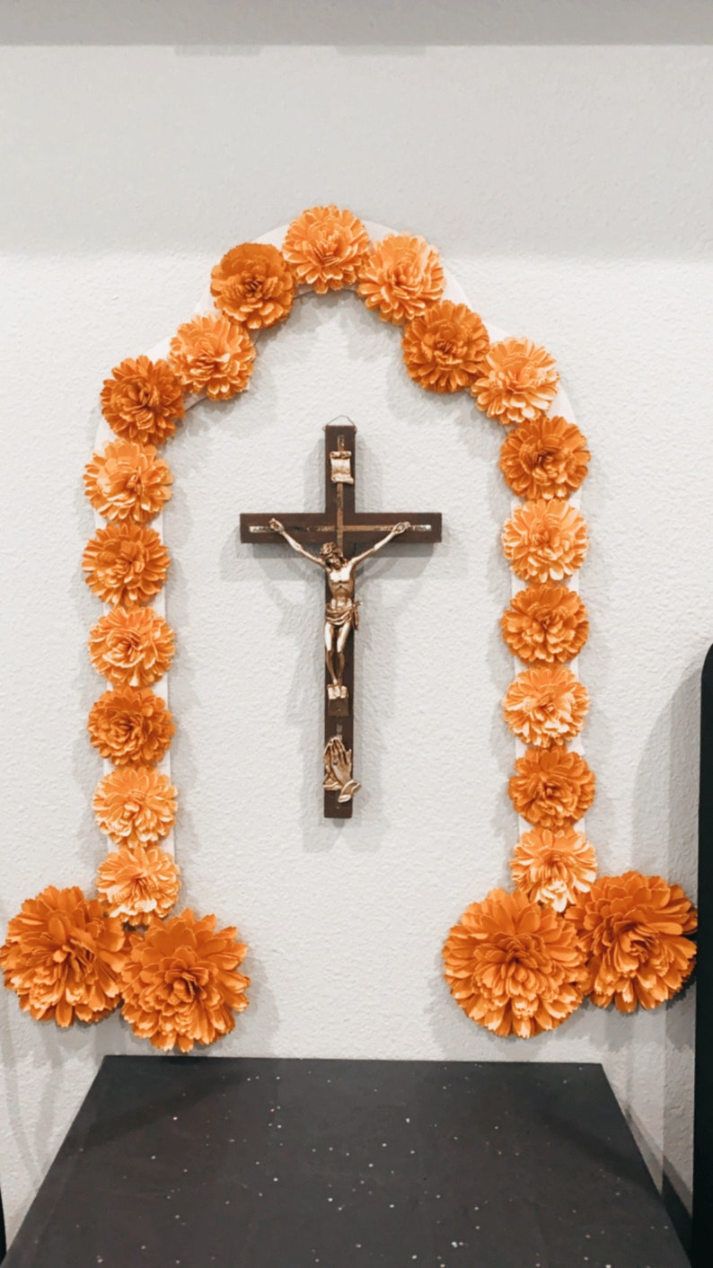 Peut inclure: Un mur blanc avec une croix en bois et une figurine dor&eacute;e de J&eacute;sus-Christ. La croix est entour&eacute;e d'une guirlande de fleurs en papier orange.