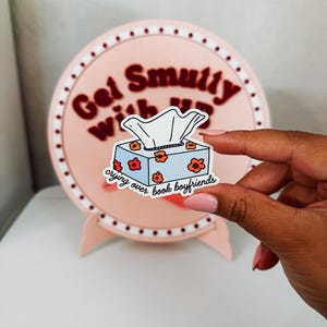 Puede incluir: Una pegatina azul claro de una caja de pañuelos con flores naranjas y el texto "crying over book boyfriends". La pegatina se sostiene frente a un letrero redondo rosa con el texto "Get Smutty With Us".