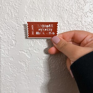 May include: A brown ticket with white text that reads "Murder Mystery Book Club" and "Admit One".