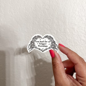 May include: A white sticker with a black skeleton hand design forming a heart shape. The sticker says "You had me at Trigger Warnings".