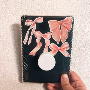 K&ouml;nnte beinhalten: Eine transparente Handyh&uuml;lle mit schwarzem Hintergrund und rosa Schleifenaufklebern. Ein wei&szlig;er PopSocket ist an der R&uuml;ckseite der H&uuml;lle befestigt.