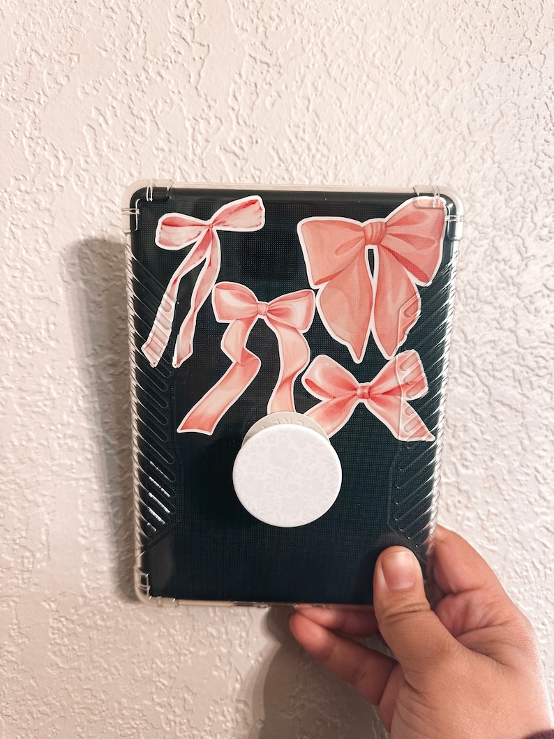 K&ouml;nnte beinhalten: Eine transparente Handyh&uuml;lle mit schwarzem Hintergrund und rosa Schleifenaufklebern. Ein wei&szlig;er PopSocket ist an der R&uuml;ckseite der H&uuml;lle befestigt.