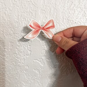 K&ouml;nnte beinhalten: Ein rosa Satin-Schleifen-Aufkleber. Die Schleife ist mit einem Knoten in der Mitte gebunden und hat eine leicht strukturierte Oberfl&auml;che.