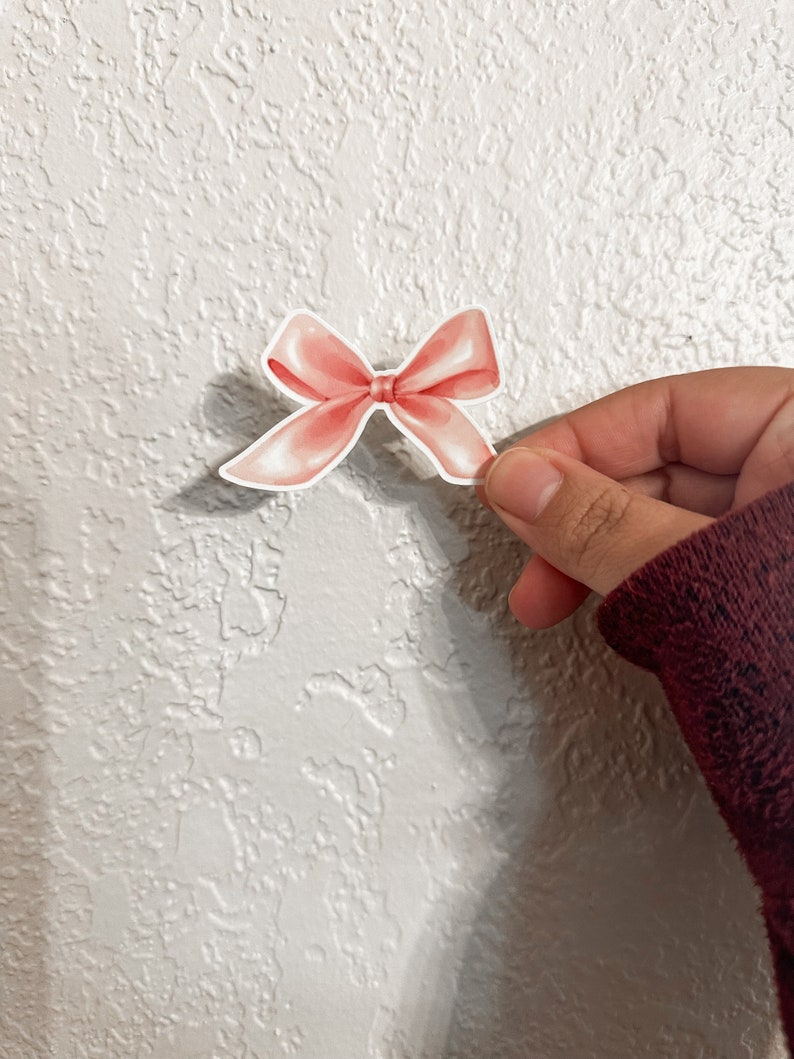 K&ouml;nnte beinhalten: Ein rosa Satin-Schleifen-Aufkleber. Die Schleife ist mit einem Knoten in der Mitte gebunden und hat eine leicht strukturierte Oberfl&auml;che.
