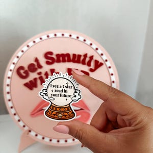 May include: A sticker featuring a crystal ball design with the text "I see a 5 star + read in your future." The sticker is held in front of a pink background with the words "Get Smutty With It" and a pair of lips.
