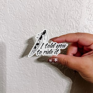 Peut inclure: Un autocollant blanc avec une illustration en noir et blanc d'une main tenant un couteau avec un motif de crâne. Le texte "I told you to ride it" est écrit sous le couteau.