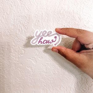 May include: A pink and white sticker with the text "yee haw" written in a rope-like font. The rope is tied into a knot at the end of the word "haw" and has a small star on it.